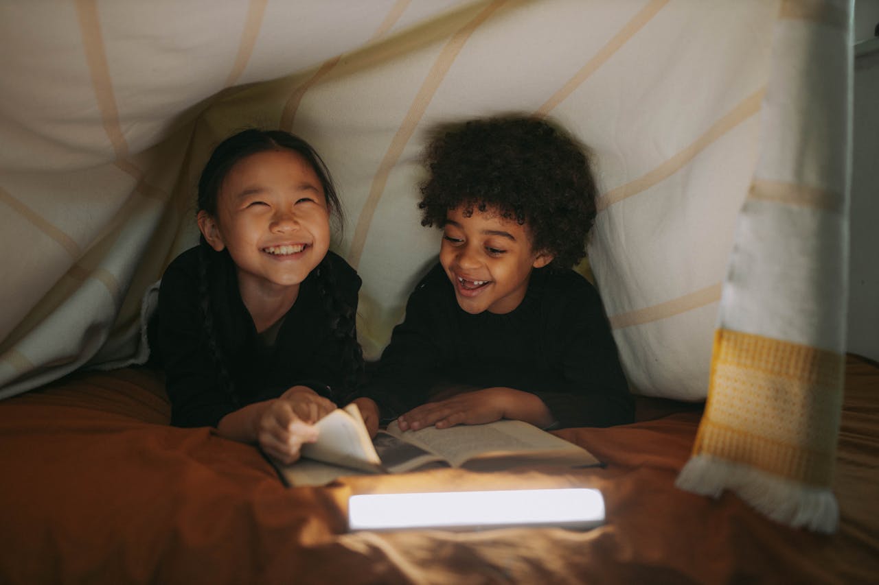 about-img Two children smiling and reading a book in a cozy blanket fort indoors.